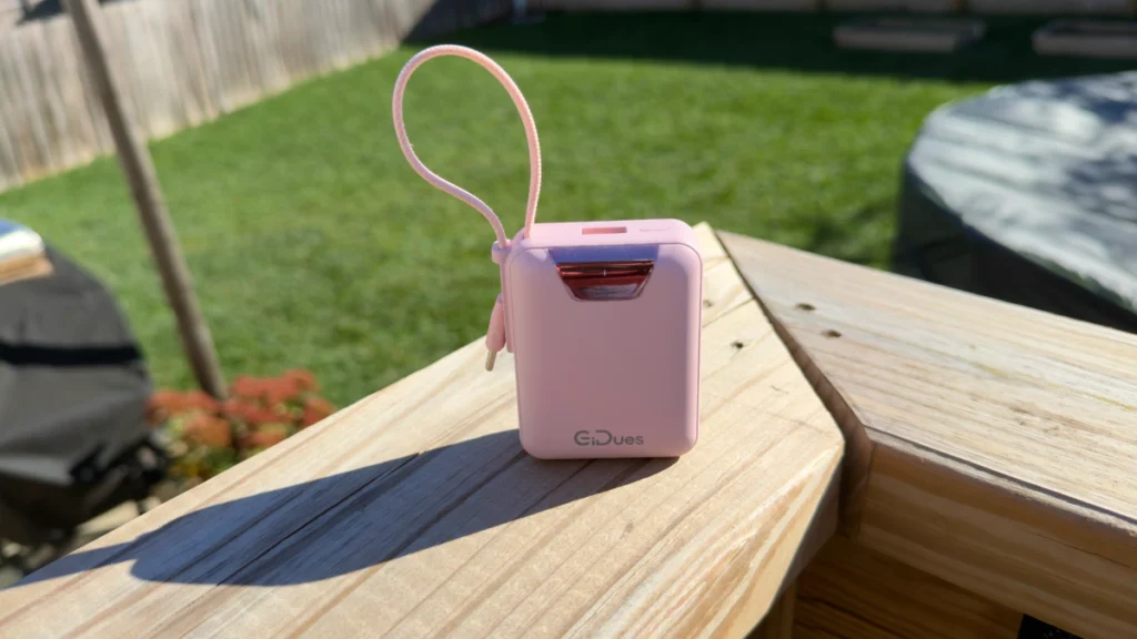 Pink Gidues M2 portable charger with its built-in USB-C cable looped on top, sitting on a deck rail in the sun