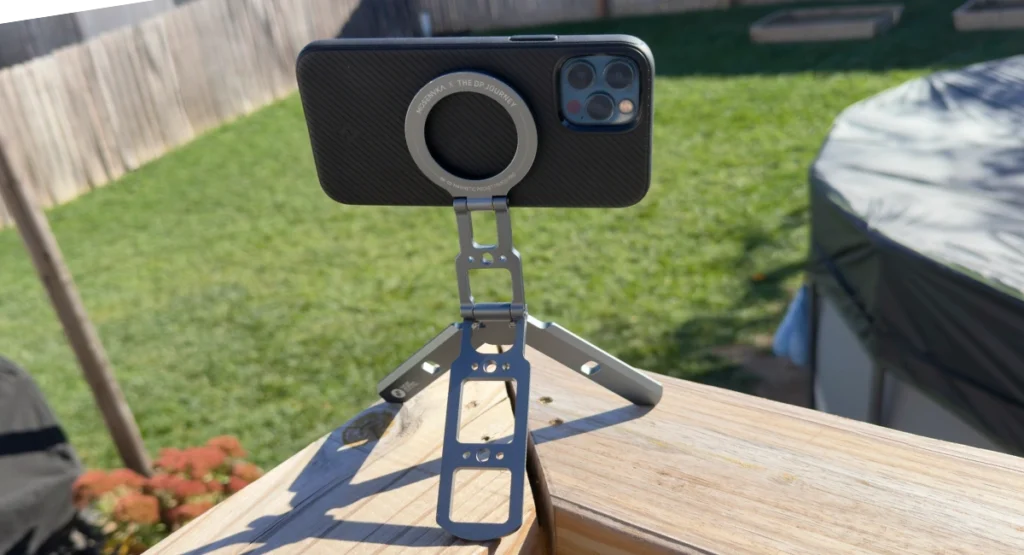 MOSENVKA magnetic phone tripod holding an iPhone 12 Pro in a black case, with its folding grey aluminum legs on a deck rail