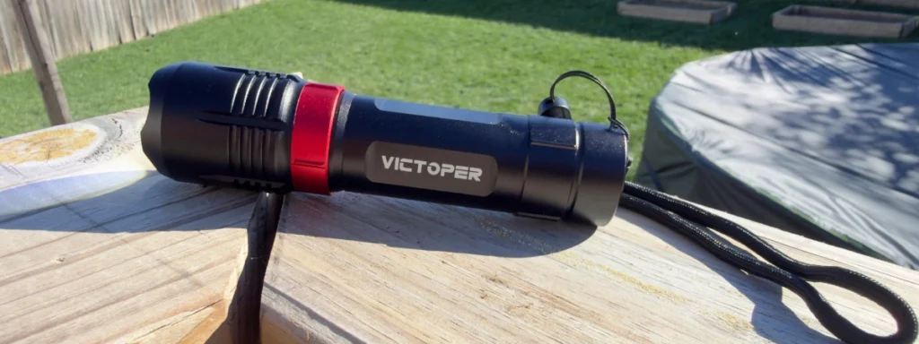 Victoper A1200T rechargeable flashlight with a red accent ring and wrist lanyard, resting on a wooden deck rail